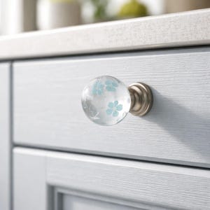 May include: A clear glass cabinet knob with light blue floral designs. The knob has a round shape and a brushed nickel base, mounted on a light blue cabinet door. The floral pattern adds a decorative touch to the hardware.