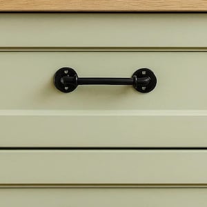May include: Close-up of a black metal drawer pull with a bar handle and circular mounting plates. The handle is mounted on a light green cabinet with a wooden top. The hardware has a rustic, industrial design.