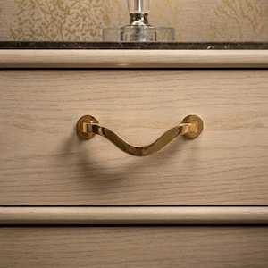 May include: Close-up of a light-colored wooden drawer with a gold-colored, curved handle. The handle is attached to the drawer with circular mounts. The drawer is part of a larger piece of furniture, possibly a dresser or nightstand.