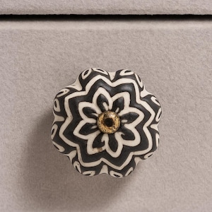May include: A decorative cabinet knob with a floral design. The knob features a black and white pattern with a gold-colored center. The knob is a unique accent for furniture.