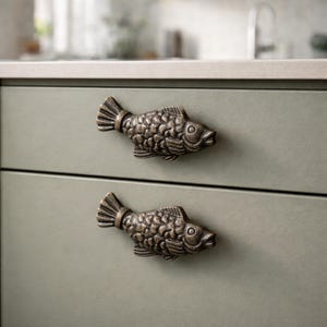 May include: Two bronze-colored fish-shaped cabinet pulls mounted on a light green cabinet. The pulls are detailed with scales and fins, adding a unique nautical touch to the kitchen or bathroom.