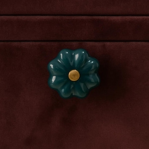 May include: A teal ceramic flower-shaped cabinet knob with a gold center. The knob is set against a dark burgundy velvet surface, creating a striking contrast. This decorative hardware adds a touch of elegance to furniture.