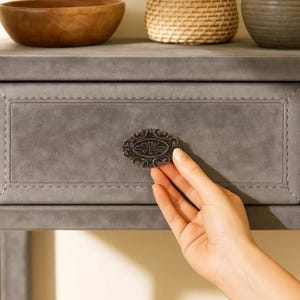 May include: Close-up of a gray drawer with a decorative, oval-shaped bronze handle. The drawer is part of a gray piece of furniture. The handle features an ornate floral design. The drawer has a stitched border.