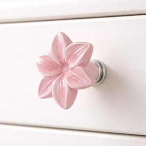May include: A pink ceramic flower-shaped drawer pull with a silver metal base. The drawer pull is mounted on a white cabinet. The flower has five petals with subtle shading, adding depth to the design. This decorative hardware is ideal for adding a floral touch to furniture.