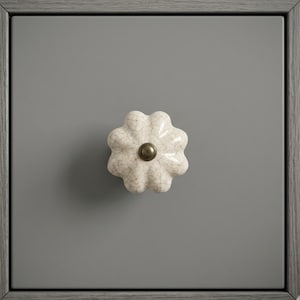 May include: A close-up of a gray cabinet door with a white, flower-shaped ceramic knob. The knob has a crackle glaze finish and a brass-colored center. The cabinet door is set within a gray wooden frame.