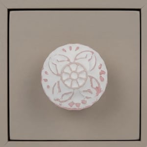 May include: Close-up of a white and pink floral design cabinet knob. The knob is round and sits on a taupe-colored drawer front. The floral design is detailed with a central circle and petal-like shapes.