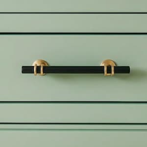 May include: A black and gold cabinet pull. The cylindrical black handle is mounted on two gold-colored circular backplates. The pull is set against a light green cabinet front with horizontal lines.