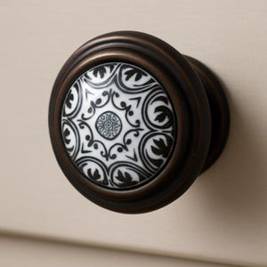 May include: A decorative cabinet knob with a dark bronze finish. The center features a white ceramic insert with a black floral pattern. The knob is round and has a classic, elegant design, suitable for furniture or cabinetry.