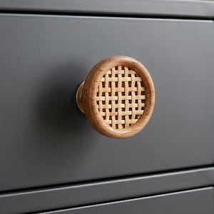 May include: Close-up of a wooden cabinet knob with a woven rattan center. The knob is round with a light brown wood frame and a natural woven pattern. The knob is mounted on a dark gray cabinet drawer.