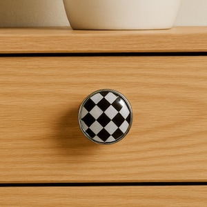 May include: A round cabinet knob with a black and white checkered pattern. The knob has a silver-colored metal rim and is mounted on a light-colored wooden drawer. The knob is a decorative hardware element.