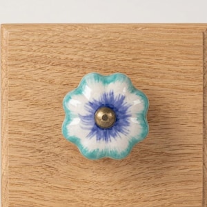 May include: A floral-shaped cabinet knob with a white, blue, and teal design and a brass-colored center. The knob is mounted on a light-colored wooden surface, showcasing a decorative hardware element.
