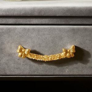May include: Close-up of a gold-colored drawer pull with a bow design on each end. The pull is adorned with a floral pattern in the center, set against a gray background. The hardware adds a touch of elegance to furniture.