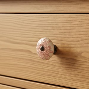 May include: A close-up of a light wood drawer with a floral ceramic knob. The knob is white with a pink floral design and a black center. The drawer has a natural wood grain.