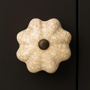 May include: Cream-colored ceramic cabinet knob with a crackle glaze finish. The knob has a flower-like shape with a dark brown center. The knob is mounted on a black surface, providing a striking contrast.