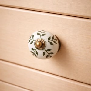 May include: A white ceramic cabinet knob with a green leaf design and a gold-colored metal base. The knob is mounted on a light-colored wooden drawer, offering a decorative accent for furniture.