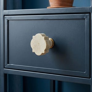 May include: Close-up of a dark blue drawer with a decorative, off-white, quatrefoil-shaped knob. The knob has a brass-colored base. The drawer is part of a dark blue metal frame, with a terracotta pot visible above.
