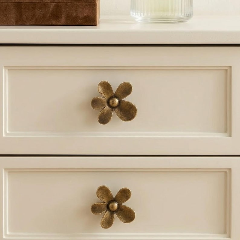 May include: Close-up of a cream-colored cabinet with two drawers, each adorned with a bronze-colored flower-shaped knob. The knobs have a central sphere and five petals. The cabinet is set against a neutral background.