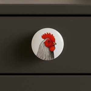 May include: A white, round cabinet knob featuring a detailed illustration of a rooster's head. The rooster has a red comb and wattle, a yellow beak, and gray and white feathers. The knob is set against a dark gray background.