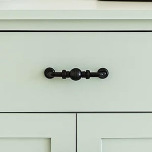 May include: Close-up of a light green cabinet drawer with a black, ornate handle. The handle is centered and has a decorative design with rounded elements. The cabinet has a simple, clean design.