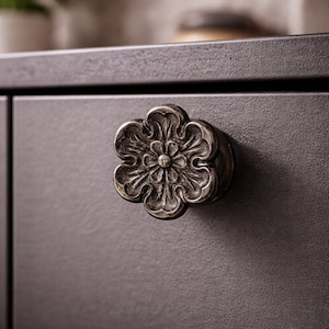 May include: Close-up of a decorative, antique-style cabinet knob. The knob is shaped like a flower with detailed petal and center designs. It is made of a metallic material with a dark, aged finish, mounted on a dark gray cabinet.