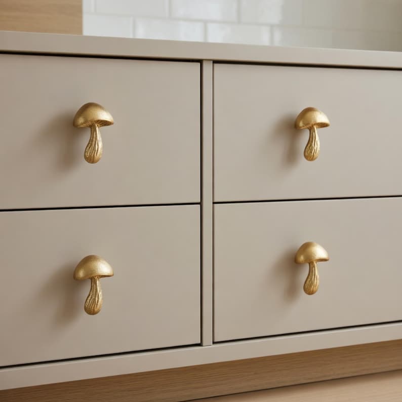 May include: Close-up of a light gray dresser with four drawers. Each drawer features a gold-colored mushroom-shaped knob. The knobs add a whimsical touch to the neutral-toned furniture, creating a unique aesthetic.