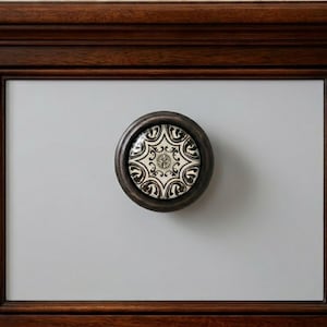 May include: Close-up of a decorative cabinet knob. The knob has a dark bronze finish and a circular ceramic insert with a black and white geometric pattern. The knob is mounted on a light gray cabinet door, framed by a dark wood border.