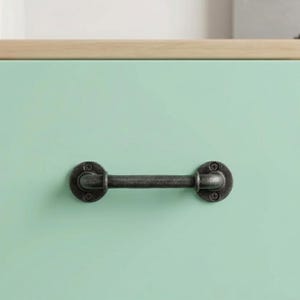 May include: A dark gray metal drawer pull with a pipe-like design. The handle is mounted on a light green surface with a wooden edge. The pull has a vintage, industrial aesthetic.
