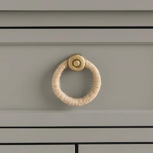 May include: Close-up of a gray cabinet drawer with a brass ring pull. The ring is wrapped in natural-colored twine, and the hardware has a circular design. The cabinet has a simple, modern design.