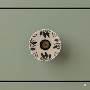 May include: A round, cream-colored cabinet knob with a black floral pattern and a brass-colored center. The knob is set against a sage green surface, suggesting a piece of furniture.