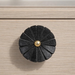 May include: A black, round cabinet knob with a textured, fan-like design. The knob features a gold-colored, spherical center. The knob is mounted on a light-colored wooden surface, providing a contrast in color and texture.