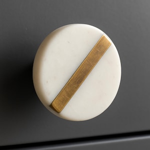May include: A round white marble cabinet knob with a diagonal brass bar. The knob is set against a dark gray background. This decorative hardware adds a touch of elegance to any furniture piece.