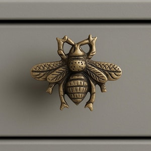 May include: A detailed, antique brass bee-shaped cabinet knob. The knob is mounted on a gray surface. The bee has textured wings, a segmented body, and intricate detailing, adding a touch of nature-inspired elegance to furniture.