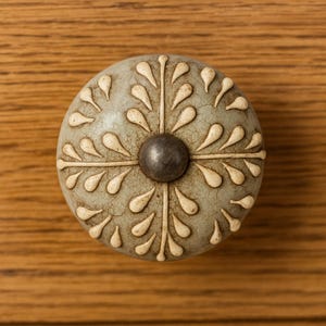May include: A round, decorative cabinet knob with a distressed, light gray finish. The knob features a central, dark gray metal accent and a pattern of off-white teardrop shapes radiating outwards. The knob is set against a wooden surface.
