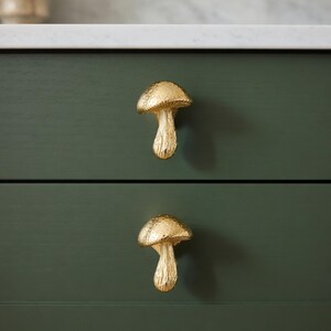 May include: Two gold-colored mushroom-shaped drawer pulls are mounted on a dark green cabinet. The mushroom caps have a textured surface, and the stems are smooth. The cabinet has a white countertop.