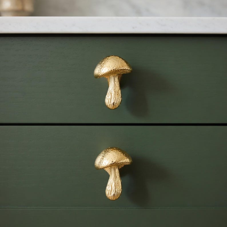 May include: Two gold-colored mushroom-shaped drawer pulls are mounted on a dark green cabinet. The mushroom caps have a textured surface, and the stems are smooth. The cabinet has a white countertop.