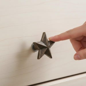 May include: A bronze-colored star-shaped cabinet knob is mounted on a light-colored wooden surface. The five-pointed star has a textured surface. A finger is touching the star knob. The knob is a decorative element for furniture.