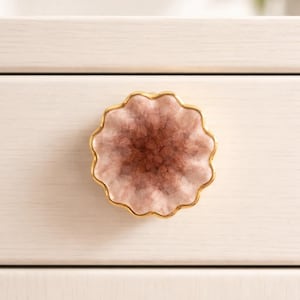 May include: A decorative cabinet knob with a scalloped edge and a gold-colored rim. The center of the knob has a pink and brown, geode-like appearance. The knob is mounted on a white cabinet drawer.