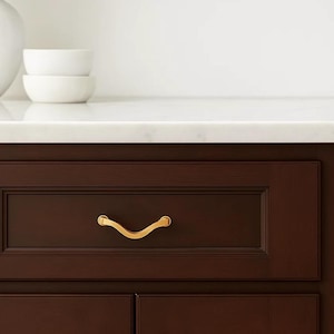 May include: A close-up of a dark brown cabinet drawer with a gold-colored, curved handle. The drawer is part of a larger cabinet with a white countertop and two white bowls in the background. The handle is approximately 5 inches long.