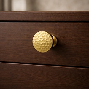 May include: A gold-colored, hammered-texture cabinet knob is attached to a dark brown wooden drawer. The knob is round with a textured surface and a small, cylindrical base. The wood grain is visible on the drawer front.