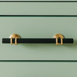 May include: A black cabinet pull with gold-colored accents. The pull is a horizontal bar with circular gold-colored mounts on each end. The cabinet door is a light green color.