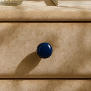 May include: A close-up of a tan, textured surface with a single, round, navy blue cabinet knob. The knob is centered and casts a shadow to the left. The background is a soft, neutral color.