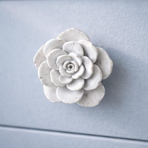 May include: A white, ceramic flower-shaped cabinet knob. The knob is mounted on a light blue surface. The flower has multiple layers of petals and a detailed center, adding a decorative touch to furniture.