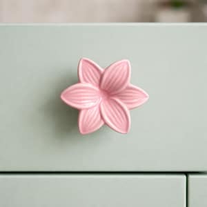 May include: A pink ceramic flower-shaped cabinet knob. The knob has six petals with subtle veining and is mounted on a light green cabinet door. The knob is a decorative accent for furniture.