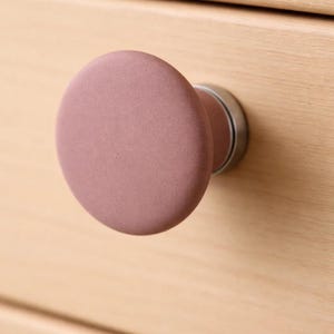 May include: A close-up of a mauve-colored cabinet knob with a rounded, oval shape. The knob is mounted on a light-colored wooden cabinet. The knob has a matte finish and a silver-colored metal base.