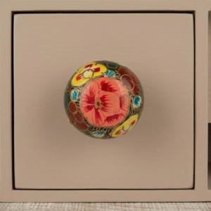 May include: A decorative cabinet knob with a floral design. The knob is round and features a central red flower surrounded by yellow, blue, and green accents. The knob is set against a light beige cabinet door.