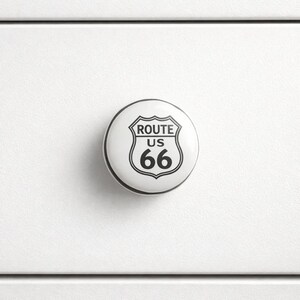 May include: A white cabinet knob with a Route 66 design. The knob is round with a black and white graphic of the iconic US Route 66 shield. The text "ROUTE US 66" is displayed in black.