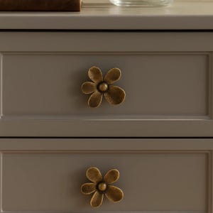 May include: Close-up of a gray cabinet with two drawers, each adorned with a bronze-colored flower-shaped knob. The knobs have a vintage, antique appearance, adding a decorative touch to the furniture.