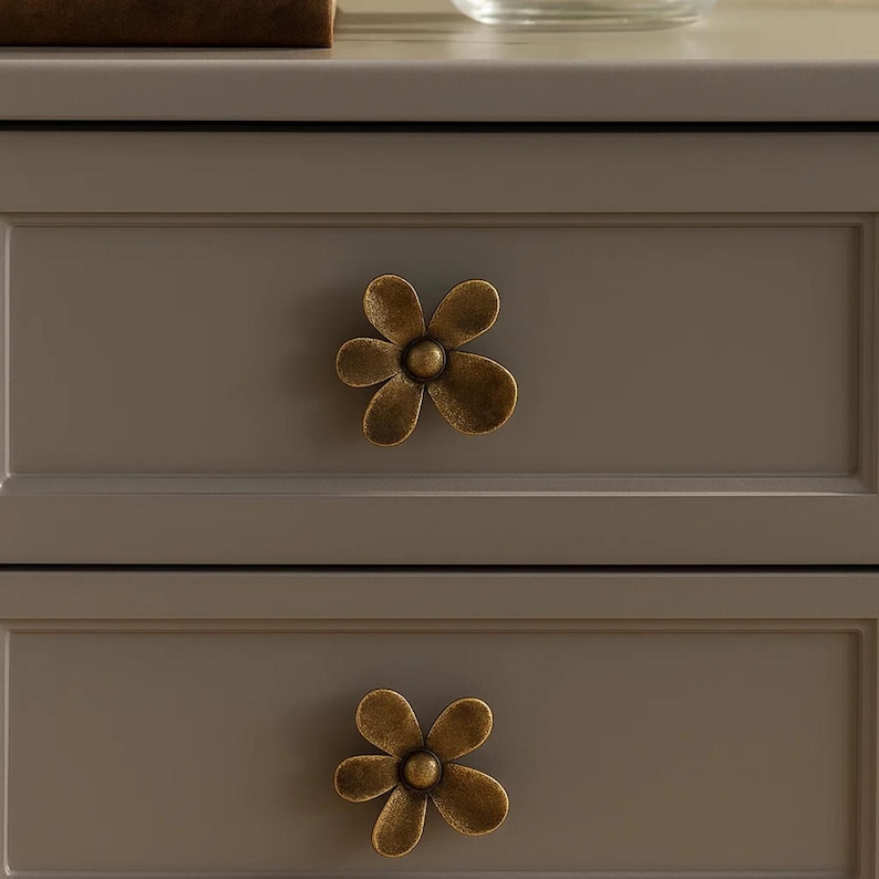 May include: Close-up of a gray cabinet with two drawers, each adorned with a bronze-colored flower-shaped knob. The knobs have a vintage, antique appearance, adding a decorative touch to the furniture.