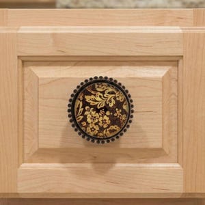 May include: A decorative cabinet knob with a floral design. The knob features a dark brown background with gold and cream-colored floral patterns. The knob is surrounded by a dark metal beaded edge and is mounted on a light-colored wooden cabinet door.