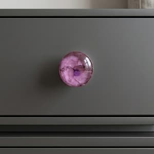 May include: A round, decorative cabinet knob with a translucent, marbled design in shades of pink and purple. The knob is mounted on a gray cabinet, offering a pop of color and a unique accent for furniture. The knob is approximately 1.5 inches in diameter.